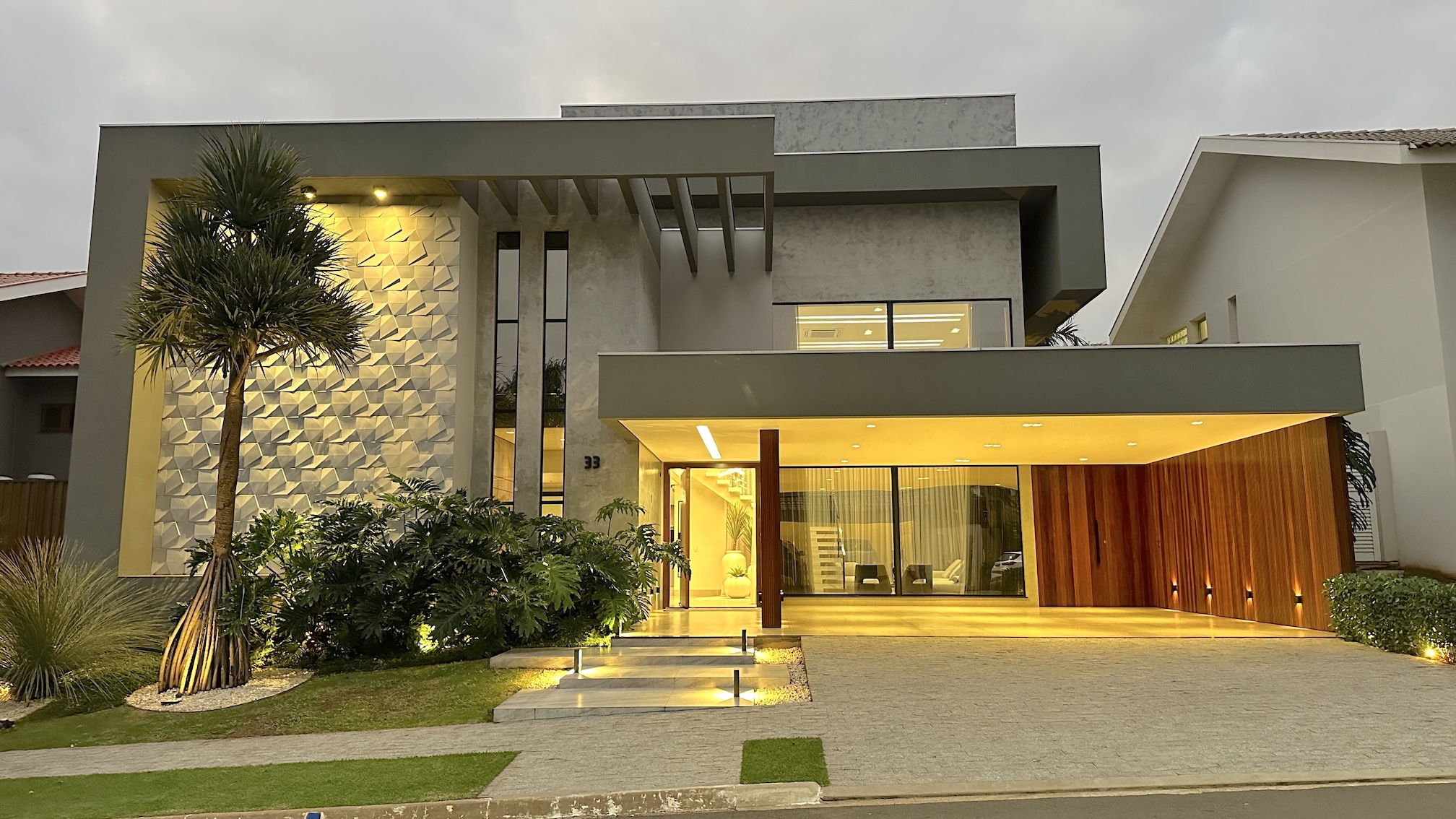 Modern luxury home exterior with large windows, stone facade, and spacious driveway at sunset.
